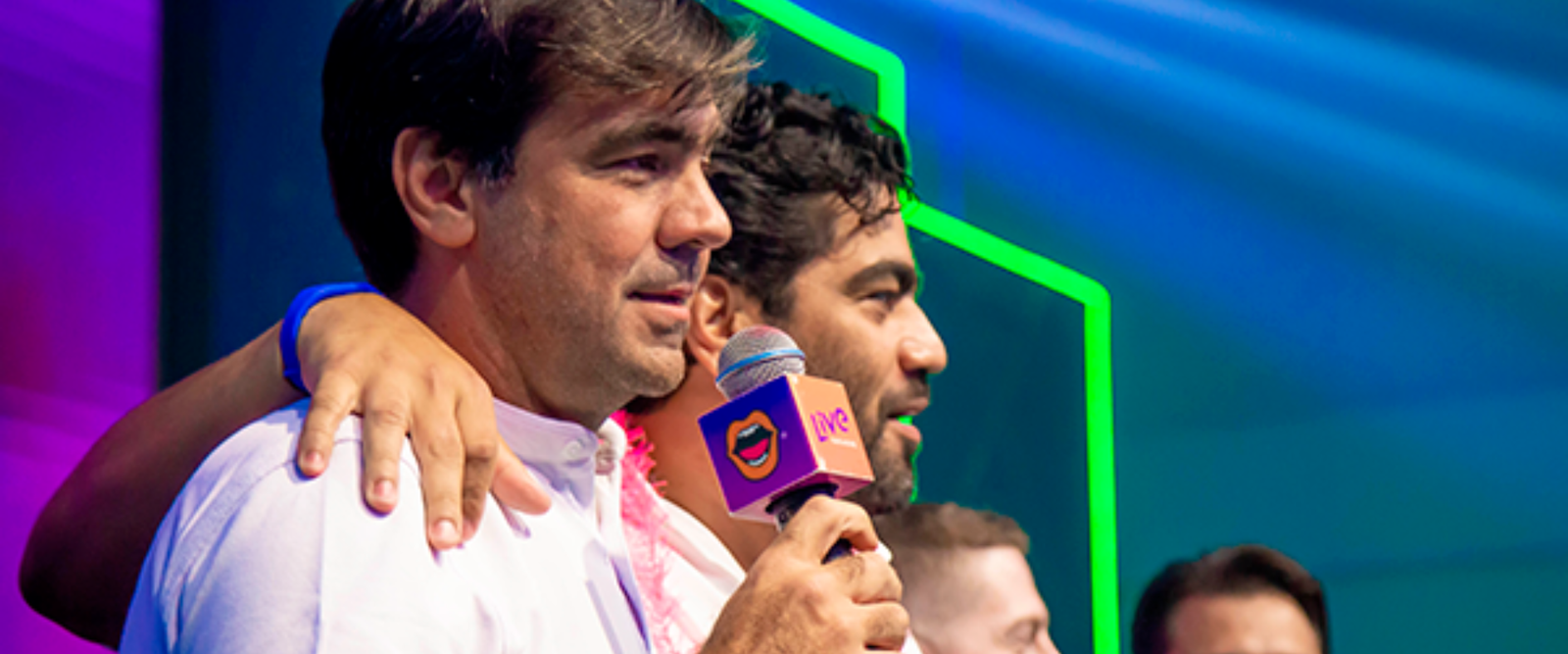 Uma banda completa se apresenta em um palco iluminado por luzes azuis e roxas. Ao fundo, um telão exibe o número "100" em branco gigante. Os músicos vestem camisas roxas e coletes pretos; há um baterista, baixista, guitarrista, tecladista e dois vocalistas.