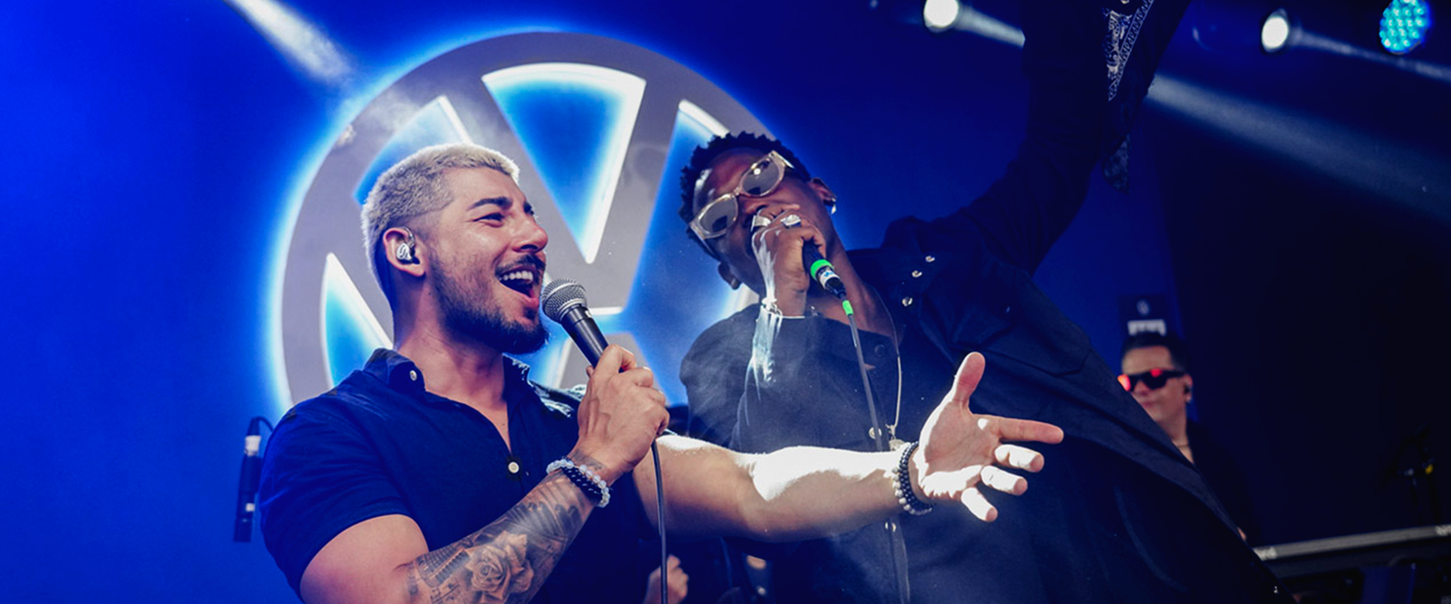 Dois homens em um palco. O homem à esquerda veste camisa jeans e segura um microfone. O homem à direita veste camiseta cinza e coloca a mão no ombro do primeiro. Ao fundo, painéis de LED com luzes roxas e neon verde.