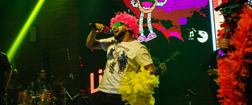 Cantora de banda de karaokê se apresenta para uma plateia engajada com mãos ao alto. A cena é iluminada por luzes de palco vibrantes, transmitindo uma atmosfera de entretenimento e festa interativa.