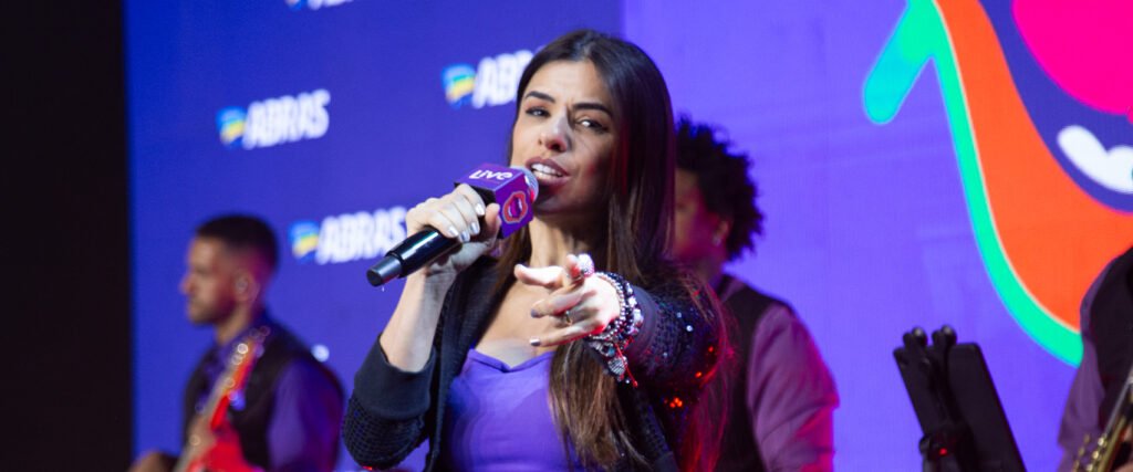 Uma cantora loira se apresenta ao vivo em um palco de karaokê, agitando uma multidão que canta junto com as mãos para o alto. A iluminação é colorida e festiva, com luzes de palco azuis e roxas.