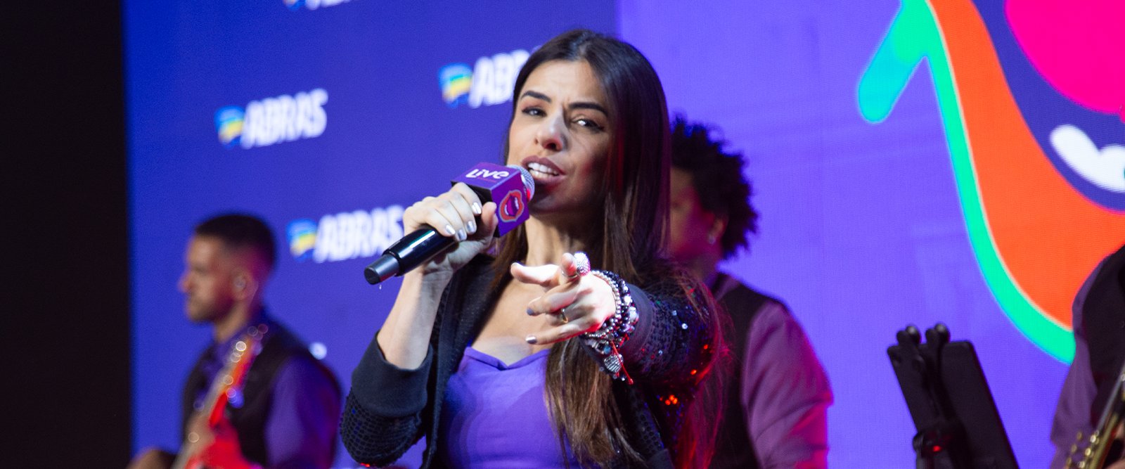 Uma cantora loira se apresenta ao vivo em um palco de karaokê, agitando uma multidão que canta junto com as mãos para o alto. A iluminação é colorida e festiva, com luzes de palco azuis e roxas.
