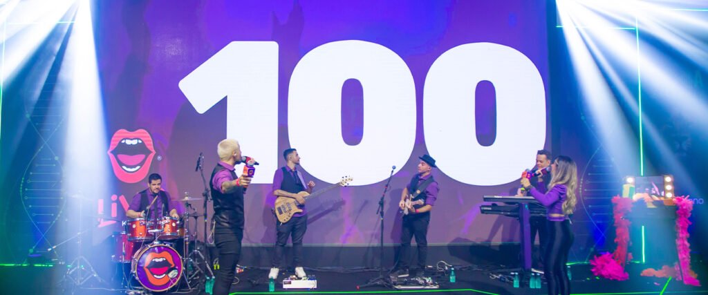Vista em plano médio de um homem de jaleco branco usando um notebook sobre uma mesa. Através de uma janela de vidro ao fundo, um menino usando fones de ouvido pretos e vermelhos e segurando um controle manual está de pé dentro de uma cabine branca.