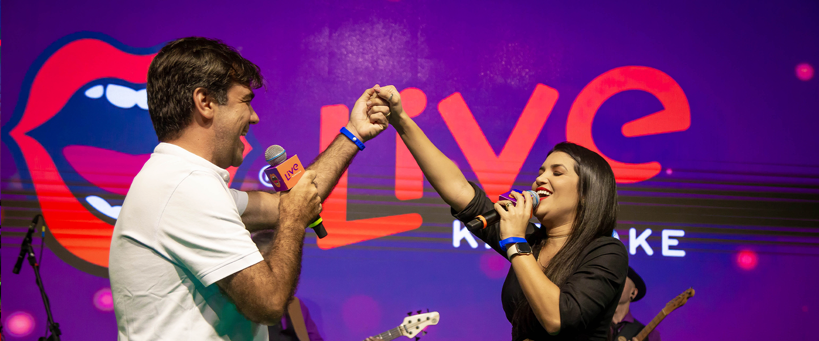 Homem e mulher cantando juntos em um palco, de mãos dadas. Eles usam microfones profissionais em frente a um telão roxo com o logotipo "Live Karaoke".