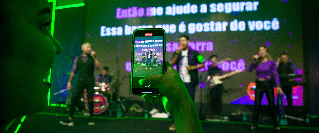 Pessoa grava com o celular uma banda se apresentando em um palco de karaokê. O telão ao fundo mostra letras de música em destaque. Vocalistas e músicos usam roupas nas cores roxo e preto sob luzes decorativas neon.