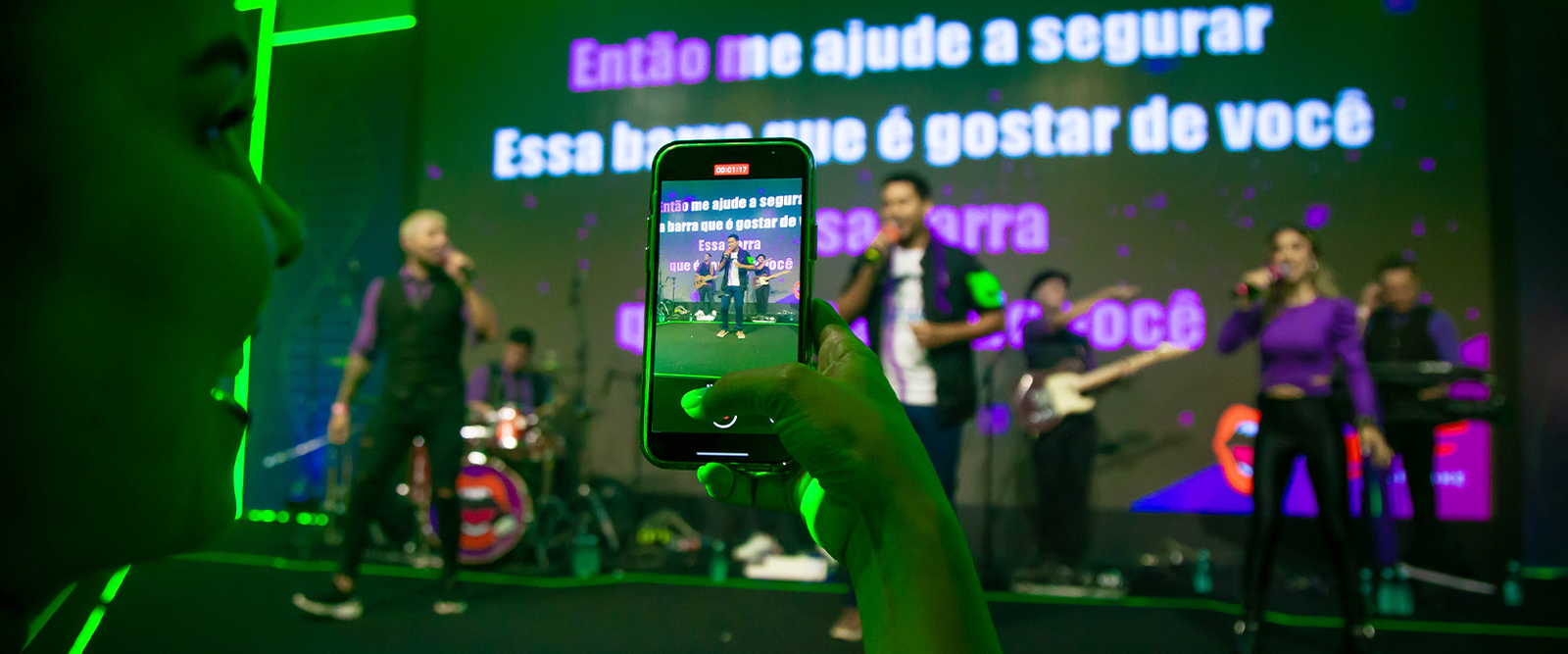 Pessoa grava com o celular uma banda se apresentando em um palco de karaokê. O telão ao fundo mostra letras de música em destaque. Vocalistas e músicos usam roupas nas cores roxo e preto sob luzes decorativas neon.