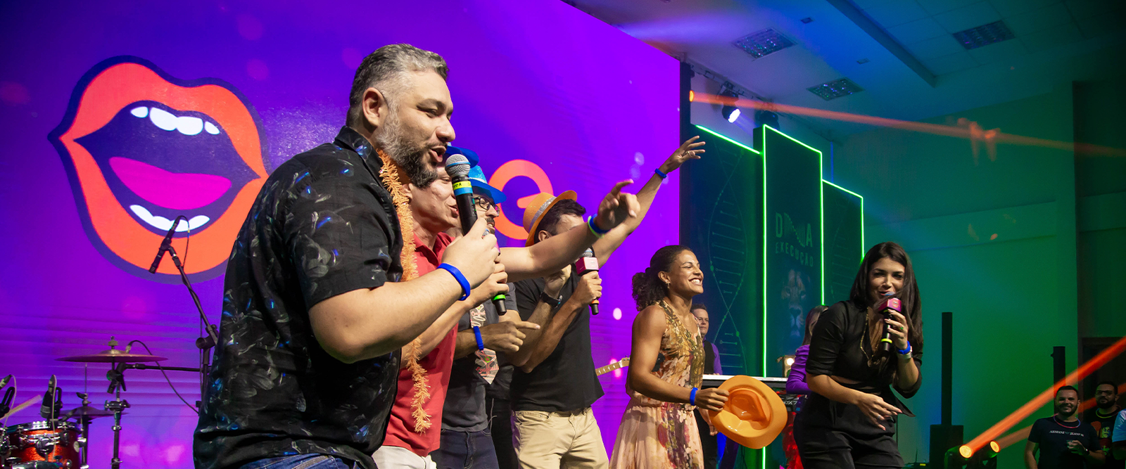 Colaboradores cantando e celebrando em um palco de evento corporativo. A imagem mostra interação, uso de adereços festivos e iluminação cênica em tons de roxo e verde.