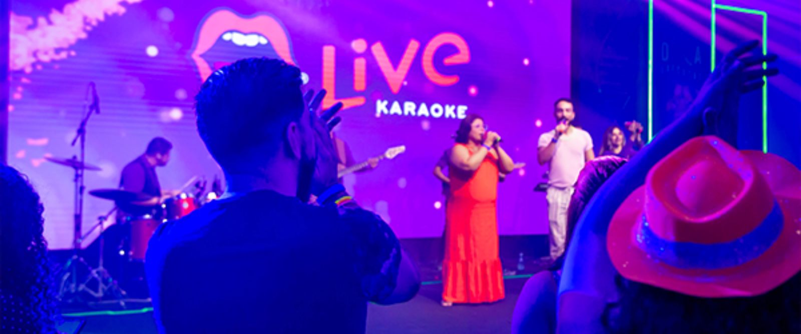 Fotografia em plano médio de um evento de "Live Karaoke". No palco, uma banda e cantores se apresentam sob luzes roxas e vibrantes, enquanto o público em primeiro plano assiste e celebra.